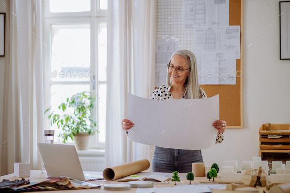 Senior Woman Architect With Model Of Houses Looking At Blueprints In 