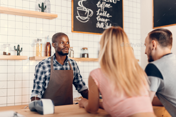 handsome african american barista talking with clients on bar counter in cafe