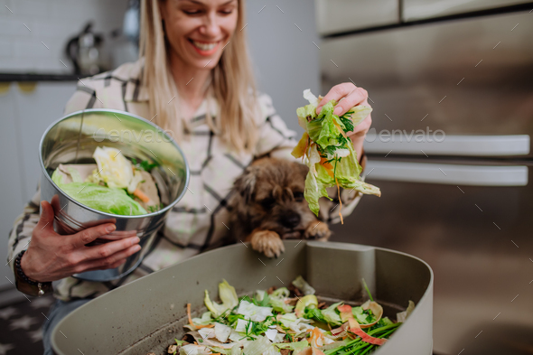 Woman throwing vegetable cuttings in a compost bucket in kitchen and feeding dog.