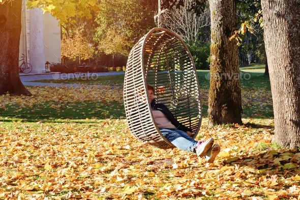 Woman in autumn park swinging on hanging rattan egg-shaped chair outdoor in midst of fallen leaves