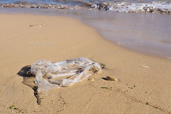 Discarded plastic bag by wave at sandy beach shore, closeup view. Global oceans pollution concept