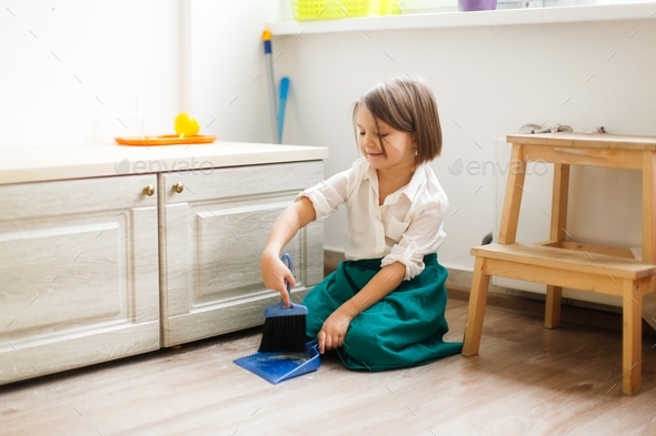 kid girl in linen apron sweeps the floor with a brush and a dustpan