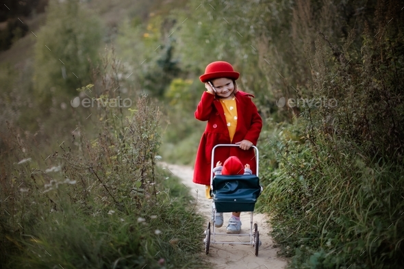 Cute caucasian girl in a red coat and hat with baby doll in stroller in park talking phone