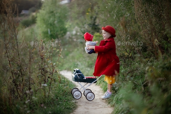 Cute caucasian girl in a red coat and hat with baby doll and doll stroller in park, child like mom