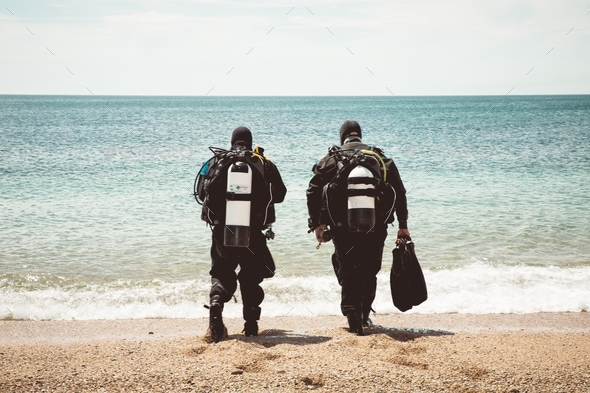Two deep sea divers wearing wetsuits and oxygen bottles walking into the ocean from a beach