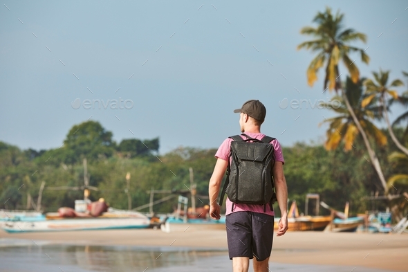 Man walking on sand beach against coastline with fishing boats. Rear view of tourist with backpack.