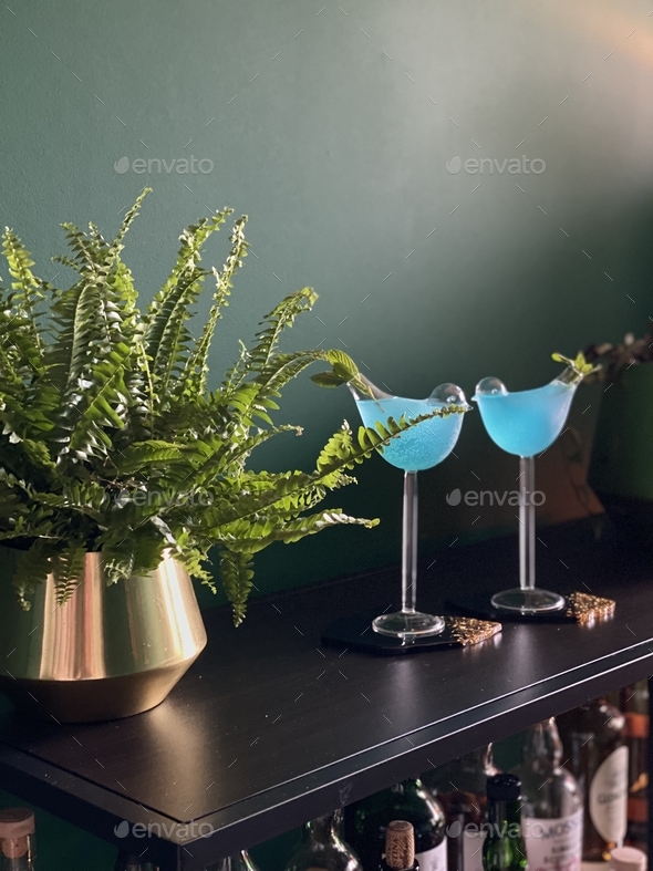 Two blue cocktails in bird shaped glasses with dark green wall at the background