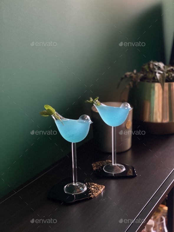 Two blue cocktails in bird shaped glasses with dark green wall at the background