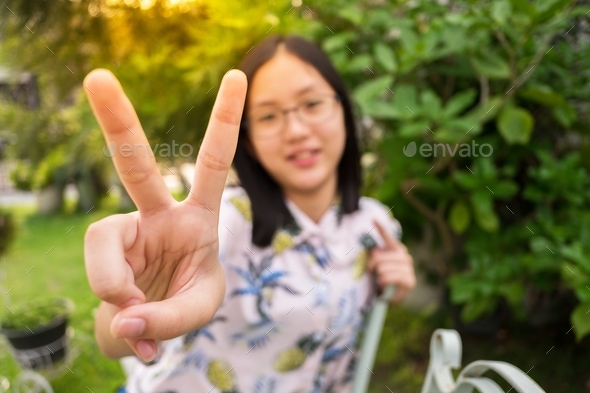 A girl with eyeglasses sitting in a garden raising her fingers in V victory