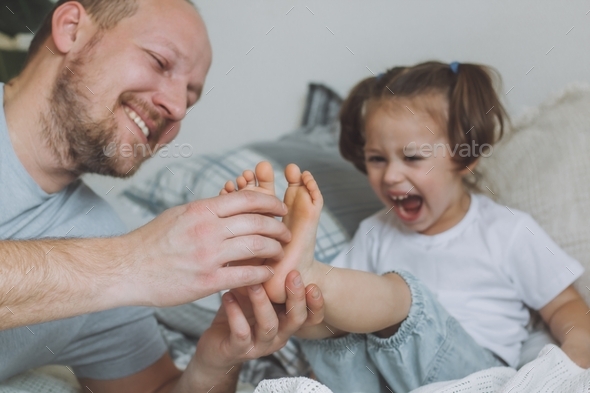 Father plays with little daughter 2-4 on bed. Dad tickles kids feet. Family having fun