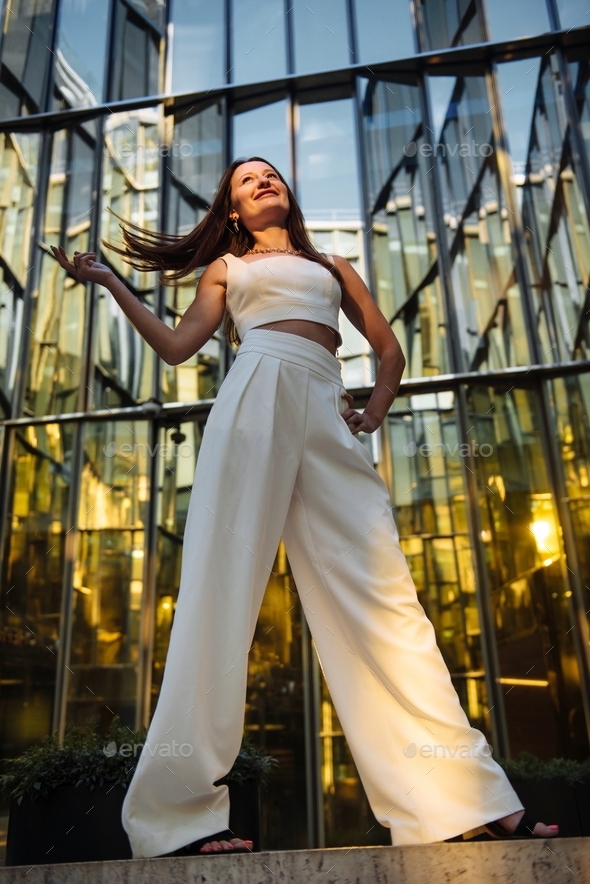 A woman in a white suit tosses her hair and smiles standing against the background of mirrored panel