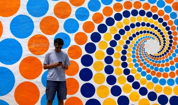 Gen Z male using technology standing on a street with a colorful art wall design in background