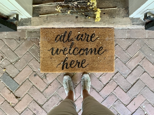 “All are welcome here” entrance rug door mat & flowers at house’s door.