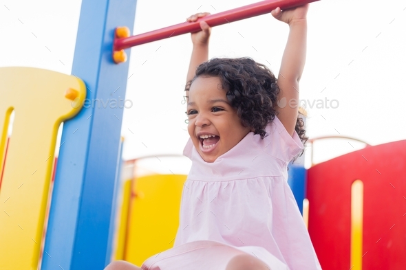 a mulatto baby with curly hair plays on a street playground. girl is hanging on a horizontal bar