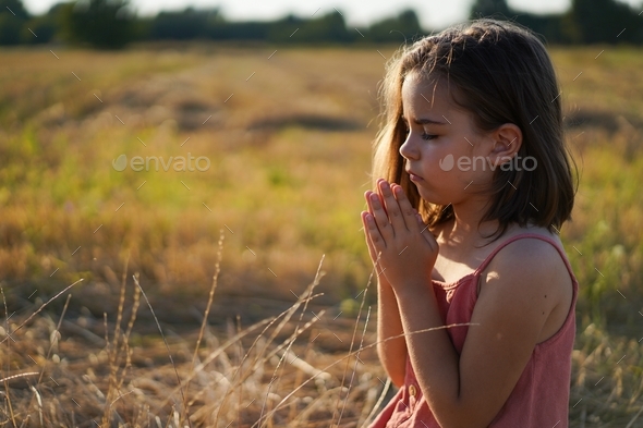 Little Girl closed her eyes, praying in a field. Hands folded in prayer.  Religion concept Stock Photo by Nastyaofly