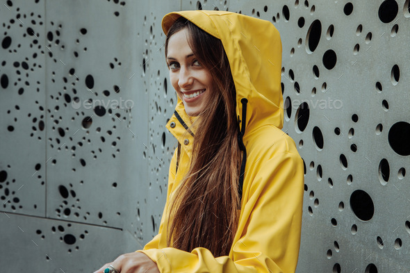 A millennial girl dressed in a yellow raincoat with a hood up looks into the camera.
