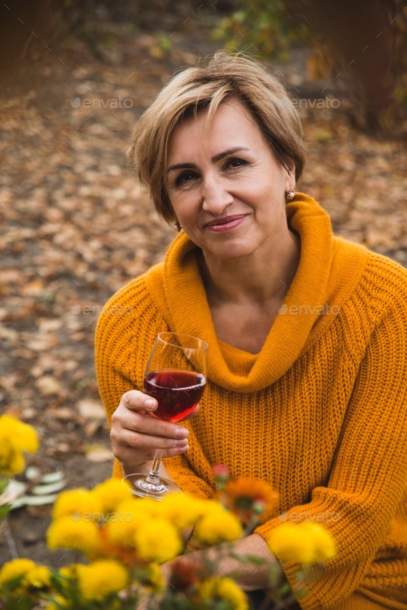 Beautiful baby boomers woman in yellow sweater holds glass of wine in backyard among autumn leaves.