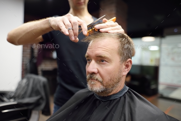 Hairdresser is cutting hair of handsome bearded mature man in salon. Stylist making hairstyle