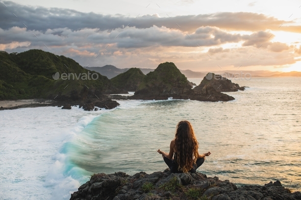Young woman practicing yoga in lotus pose at sunset with beautiful ocean and mountain view