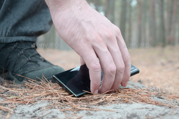 Male hand picking up lost mobile phone from a ground in autumn fir wood path