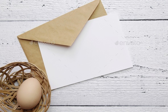 egg in a basket and envelope with empty greeting card on a white wooden table