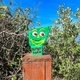 Green painted Gumby rock on wood stake in bushes under blue sky. - PhotoDune Item for Sale