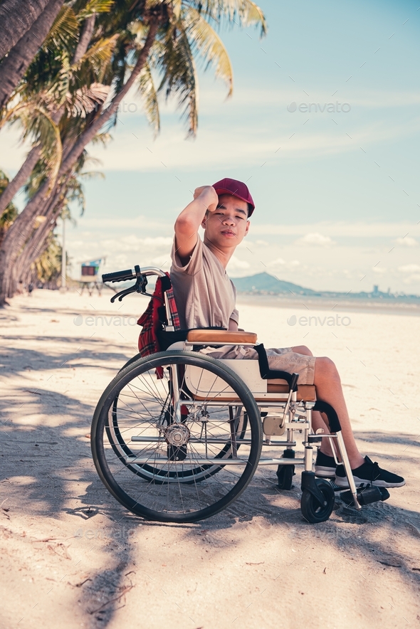Happy handicapped teenager boy in wheelchair holding red cap and smiling face,Sea beach background.