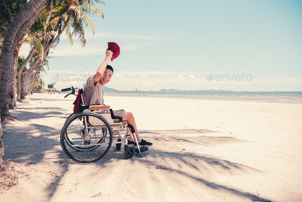 Happy handicapped teenager boy in wheelchair holding red cap and smiling face,Sea beach background.