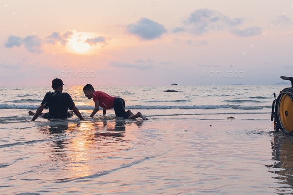 Silhouettes of disabled child and parent crawling on the sea beach at sunset and wheelchair.
