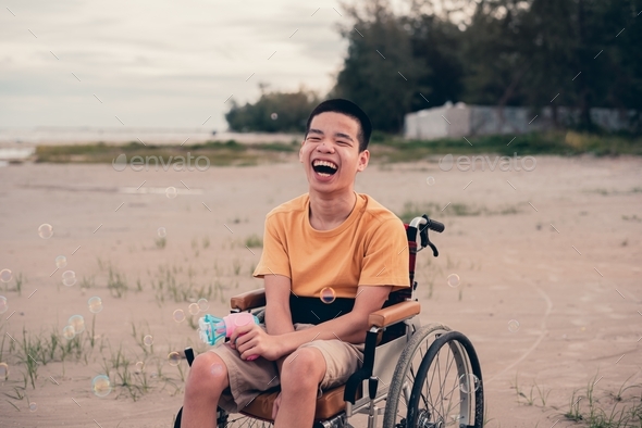 Handicapped teenager boy on wheelchair playing bubble gun on the beach.