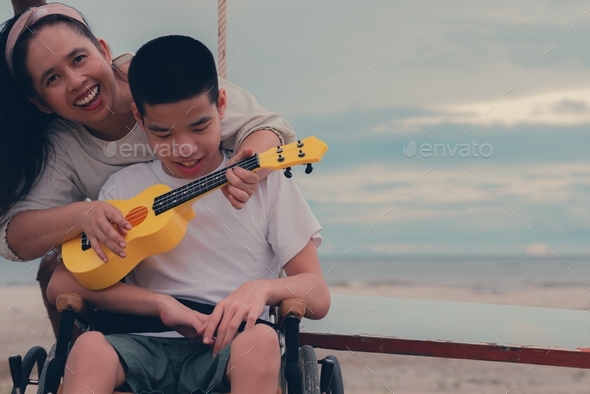 Happy disabled teenager boy on wheelchair and mother relax with music therapy on the sea beach.