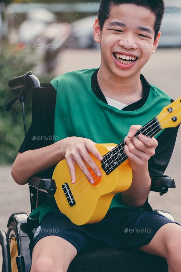 Happy handicapped teenager boy on wheelchair play yellow ukulele with smiling face.