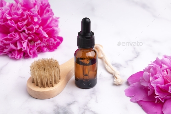 Cosmetic oil in a glass dropper bottle and massage brush on marble background with peonies