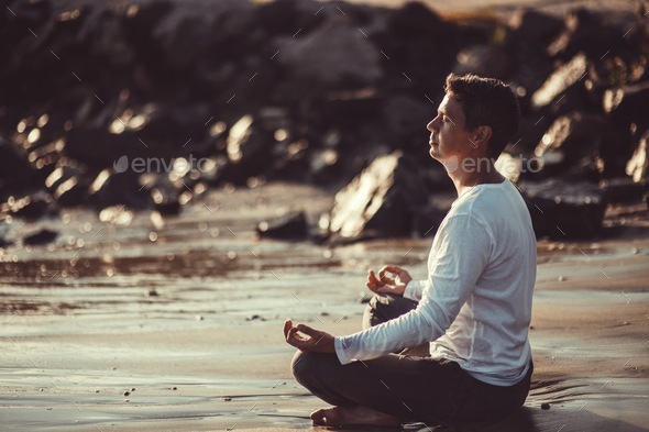 Candid authentic male millennial sitting in lotus pose on sea rocky shore greets dawn