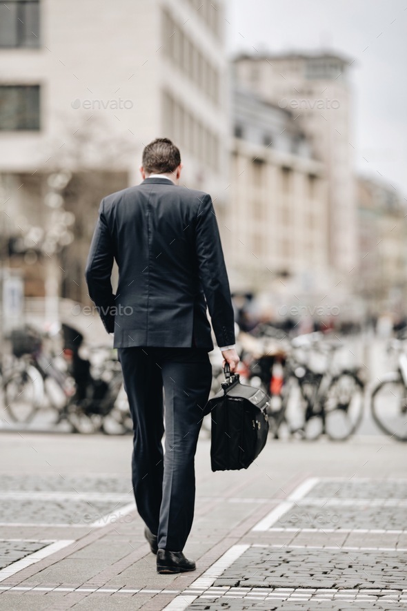 Businessman with hand in pocket from behind walking on city street to office with black briefcase