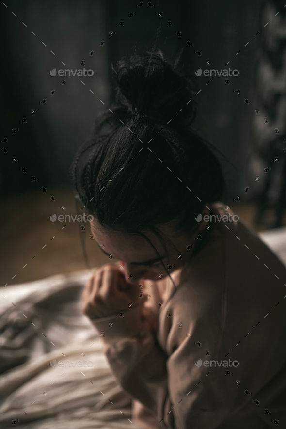 Candid sad brunette with bun of hair in beige clothes of earth tones girl sit on the bed and suffers