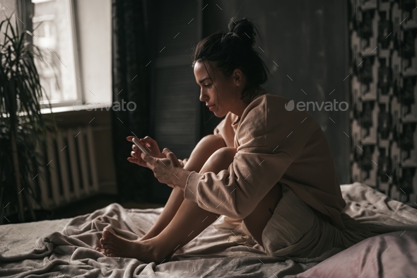 Candid sad brunette with bun of hair in beige clothes of earth tones girl sit on the bed and suffers
