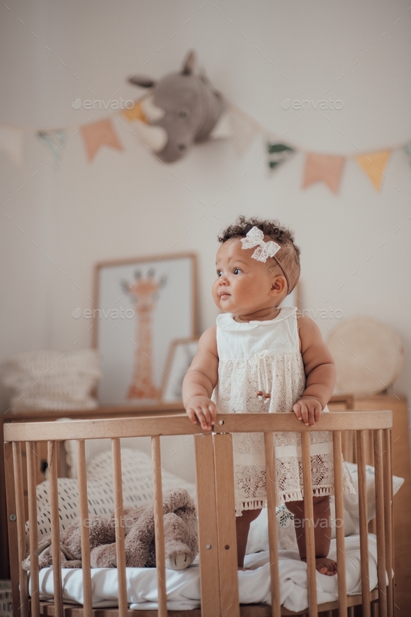 Mulatto infant girl standing in round wooden baby crib at cozy children\'s room in pastel colors