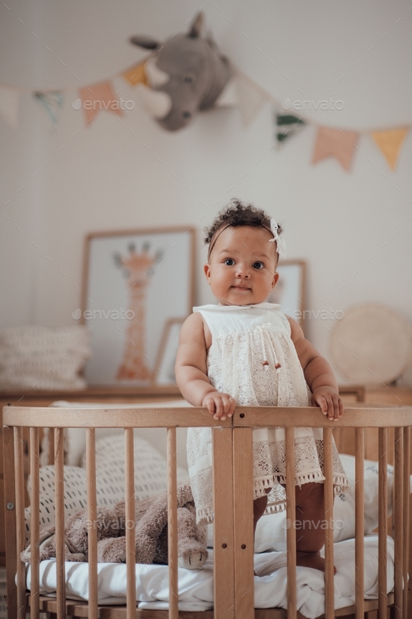 Mulatto infant girl standing in round wooden baby crib at cozy children\'s room in pastel colors