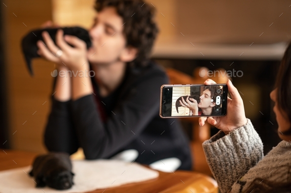 A girl shoots a video on a phone camera as a young man kisses a small black puppy of Labrador