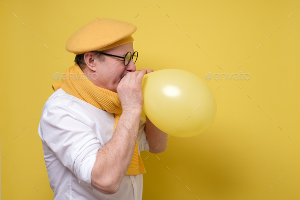 man in yellow beret and scarf getting ready to the party blowing a balloon.