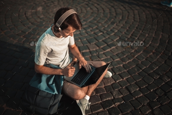 Millennial student using technology outdoor. Man using wireless headphones and laptop