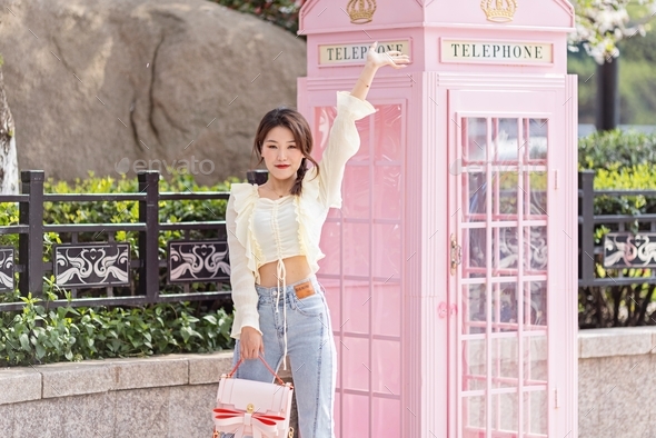 Young stylish Chinese woman standing near pink telephone booth outdoor