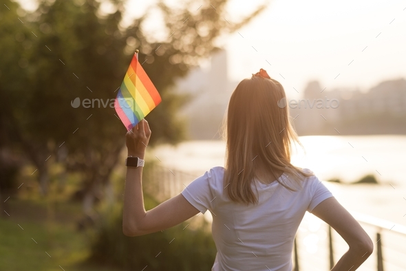 Woman in white t-shirt holding lgbtq rainbow flag outdoors on sunset. She standing from behind.