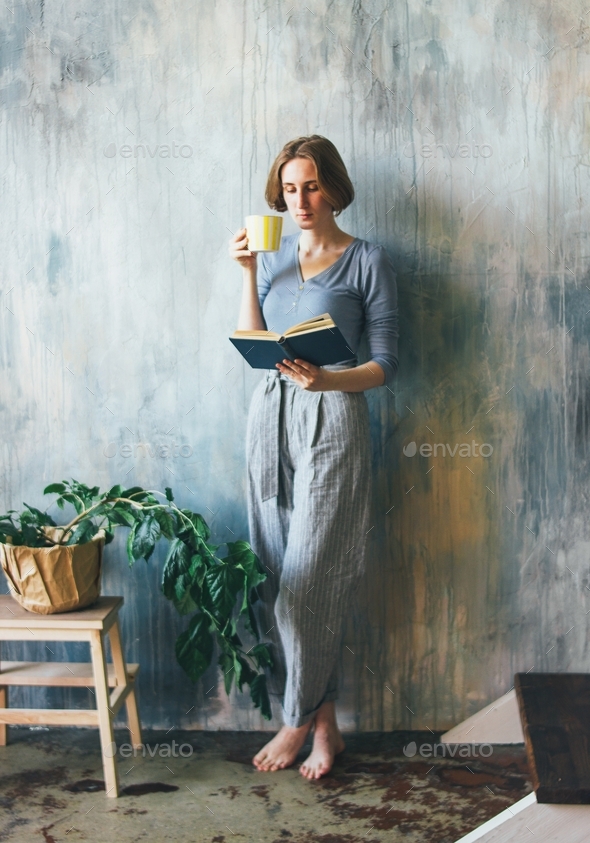 Young woman with yellow cup standing against wall and reading book in the art work studio
