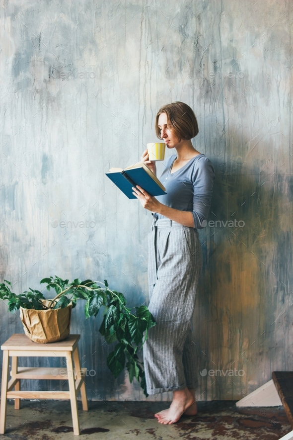 Young woman with yellow cup standing against wall and reading book in the art work studio