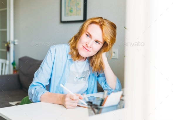 Young woman with red hair illustrator web designer draws on tablet at desk at the home office