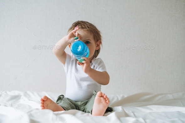 Cute happy funny baby girl in home clothes drinking water from bottle on bed at home