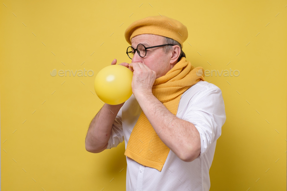man in yellow beret and scarf getting ready to the party blowing a balloon.