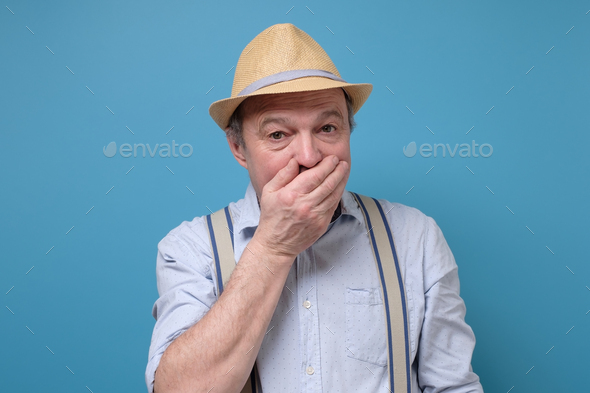 Stunned elderly man in summer hat covers mouth with hands, being surprised